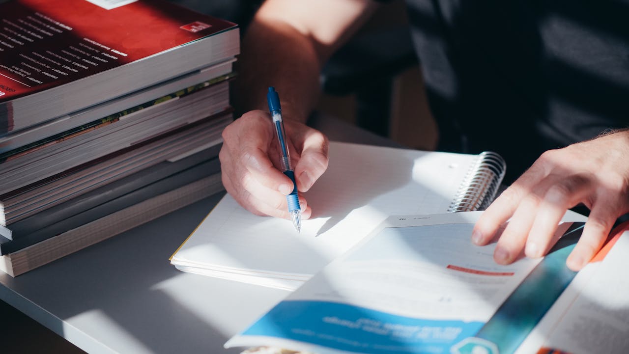A person writing notes in a notebook with a stack of textbooks nearby, captured indoors.